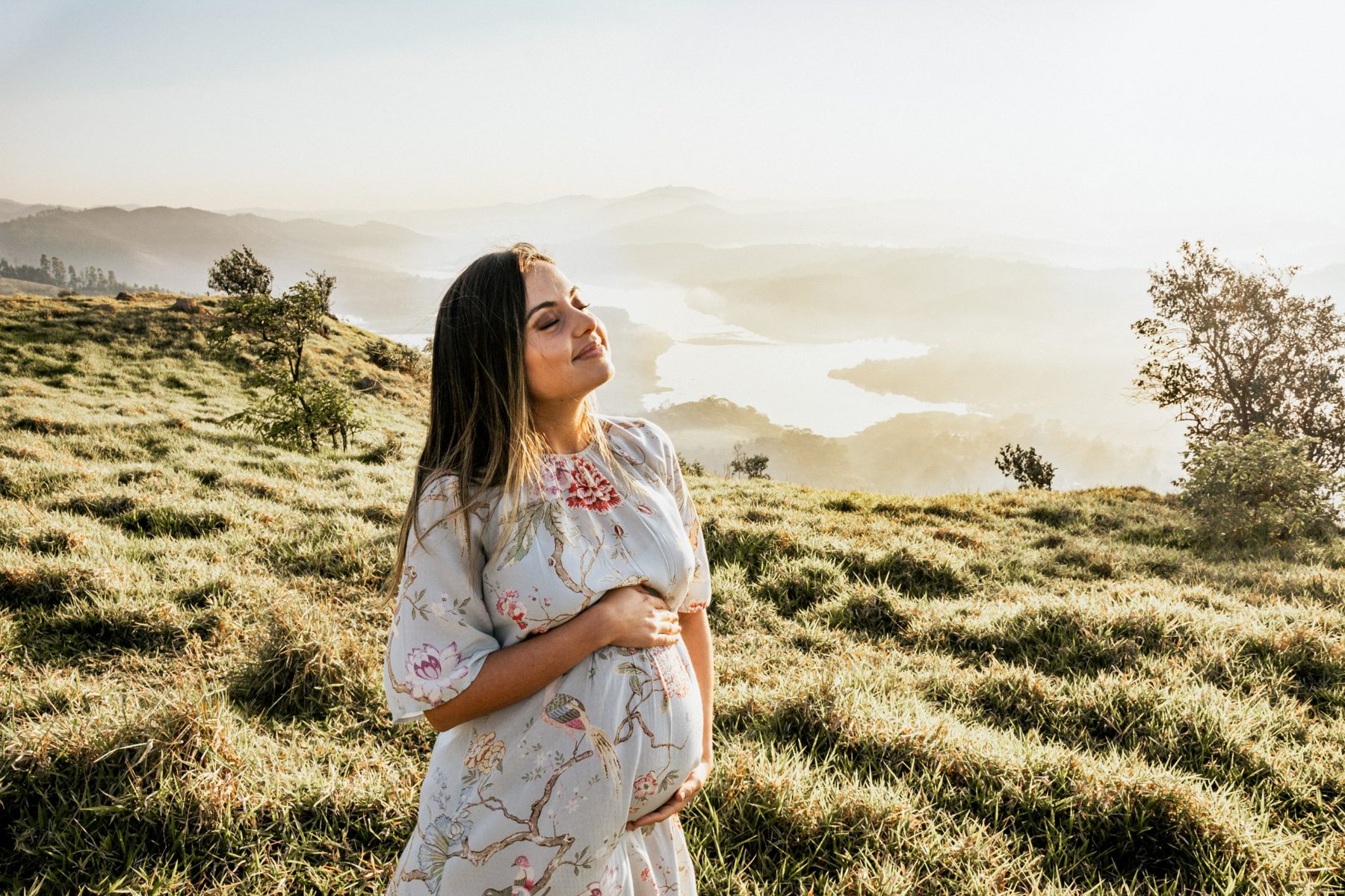 Gestante de olhos fechados no campo ao sol, com as mãos na barriga