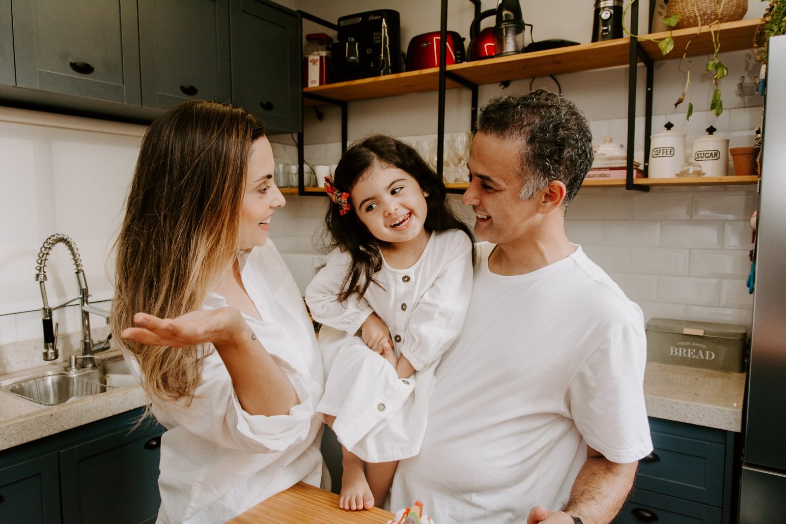 Pai segurando a filha no colo na cozinha, os três sorrindo