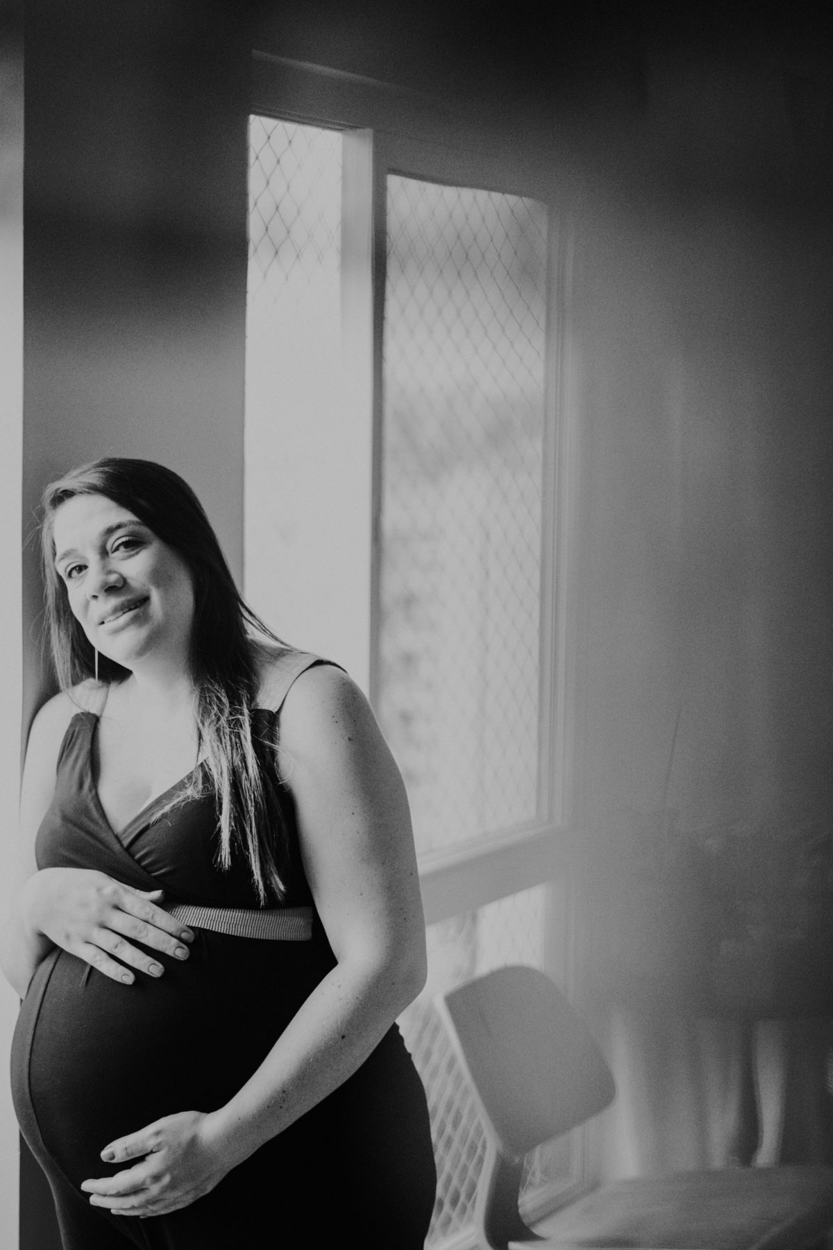 Gestante sorrindo perto da janela, em preto e branco