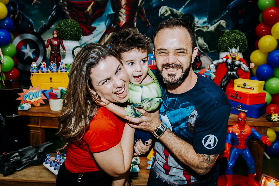 Pais abraçando o filho, rindo juntos, em frente à mesa da festa