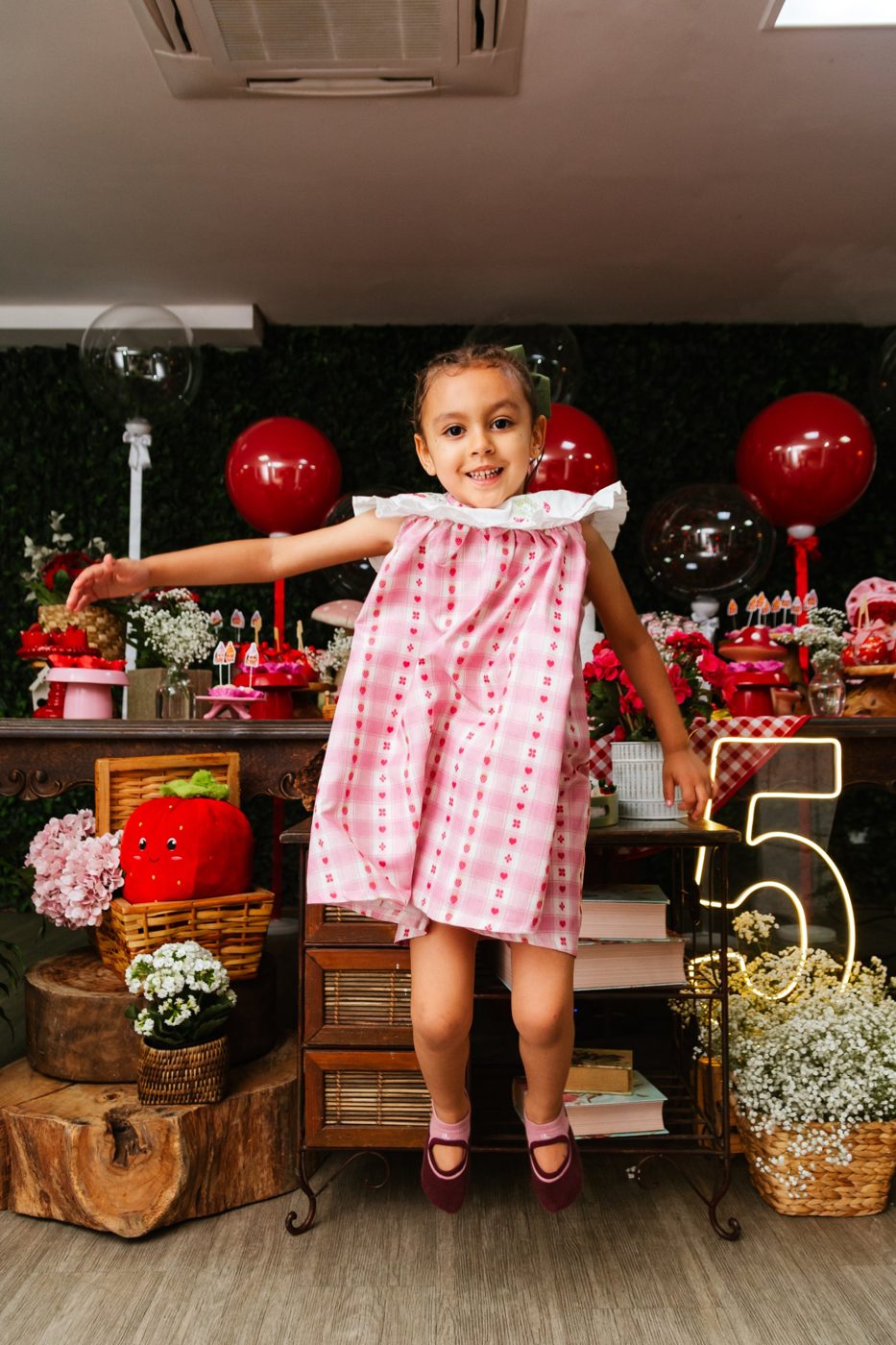 Menina de vestido xadrez rosa pulando em frente à mesa da festa